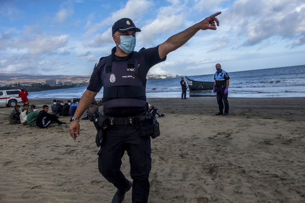 Un agente de policía da instrucciones a reporteros mientras migrantes marroquíes permanecen sentados en la playa tras su llegada a las costas de la Isla Canaria. (AP)